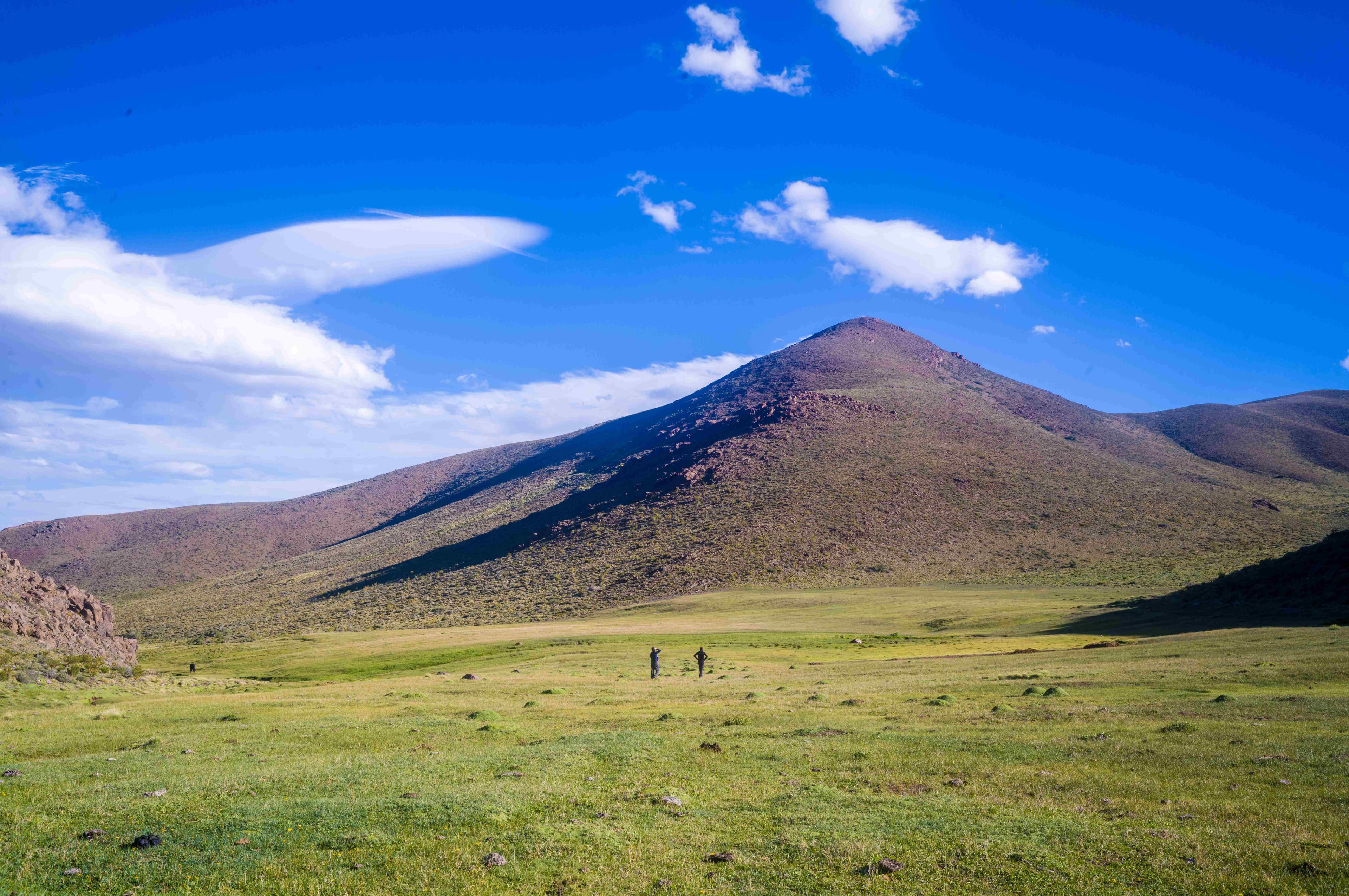 Dos caminantes en pradera verde con cerro volcánico y cielo azul